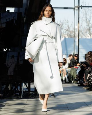 Stella McCartney model walking in the F/W 24 show wearing a white leather funnel-neck trench coat with a white bag and white pumps.