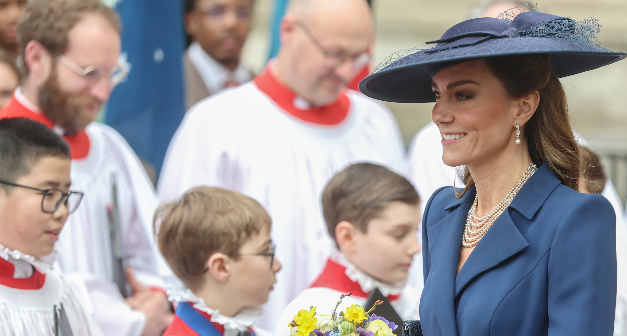Princess Kate wearing a blue coat and hat holding flowers in front of choir boys