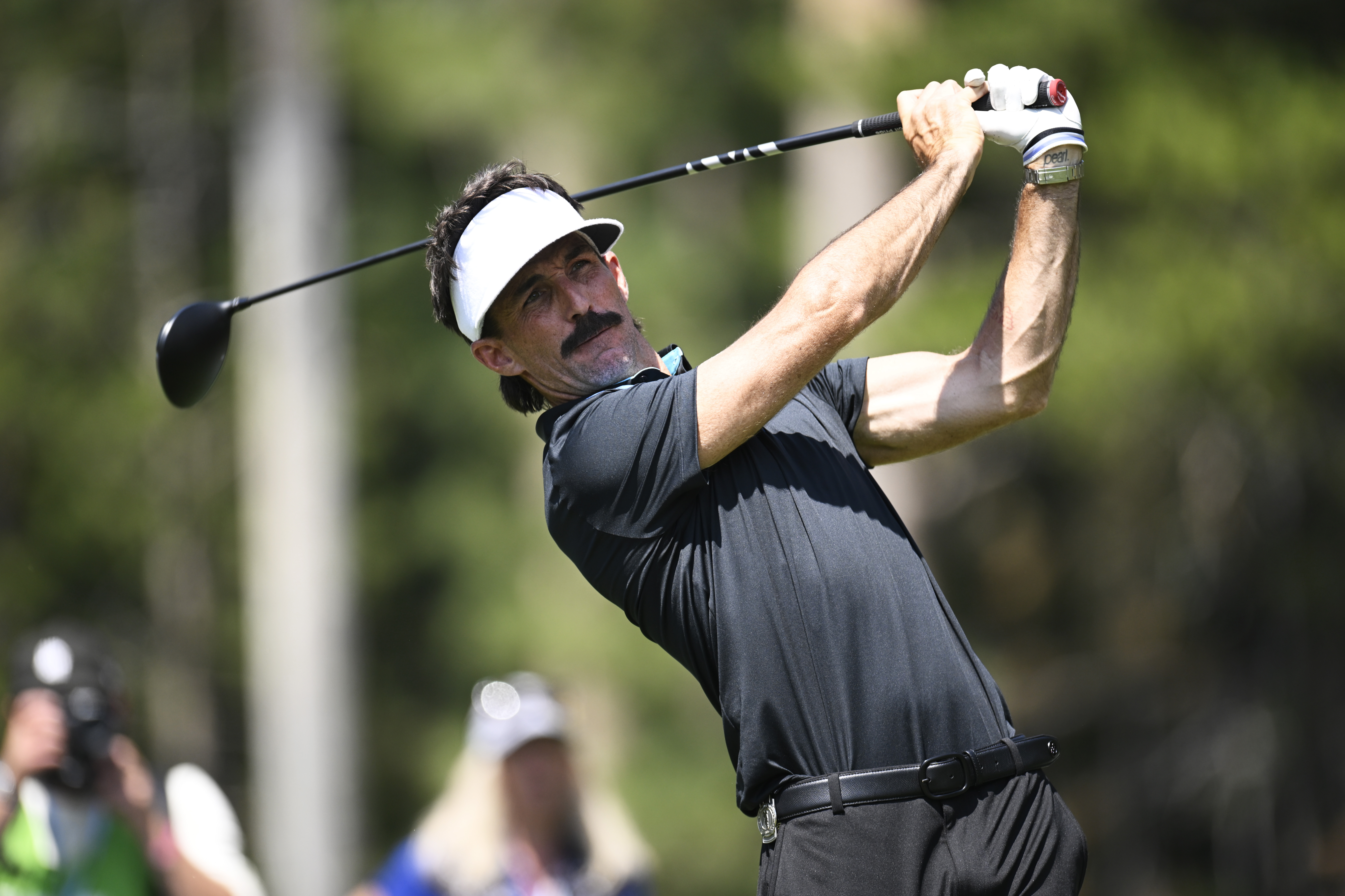 Jake Owen holds his finish on a drive while wearing black pants and a black polo with a white visor