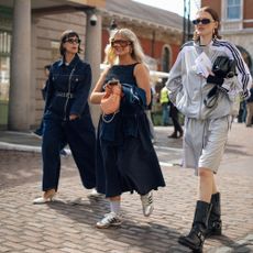 A group of three. women walking. One wears a denim jumpsuit. Another wears a denim dress and adidas. The last one wears a track jacket, shorts, and long boots. 