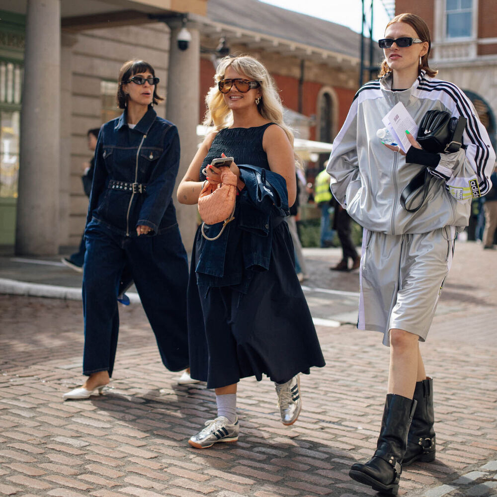 A group of three. women walking. One wears a denim jumpsuit. Another wears a denim dress and adidas. The last one wears a track jacket, shorts, and long boots.