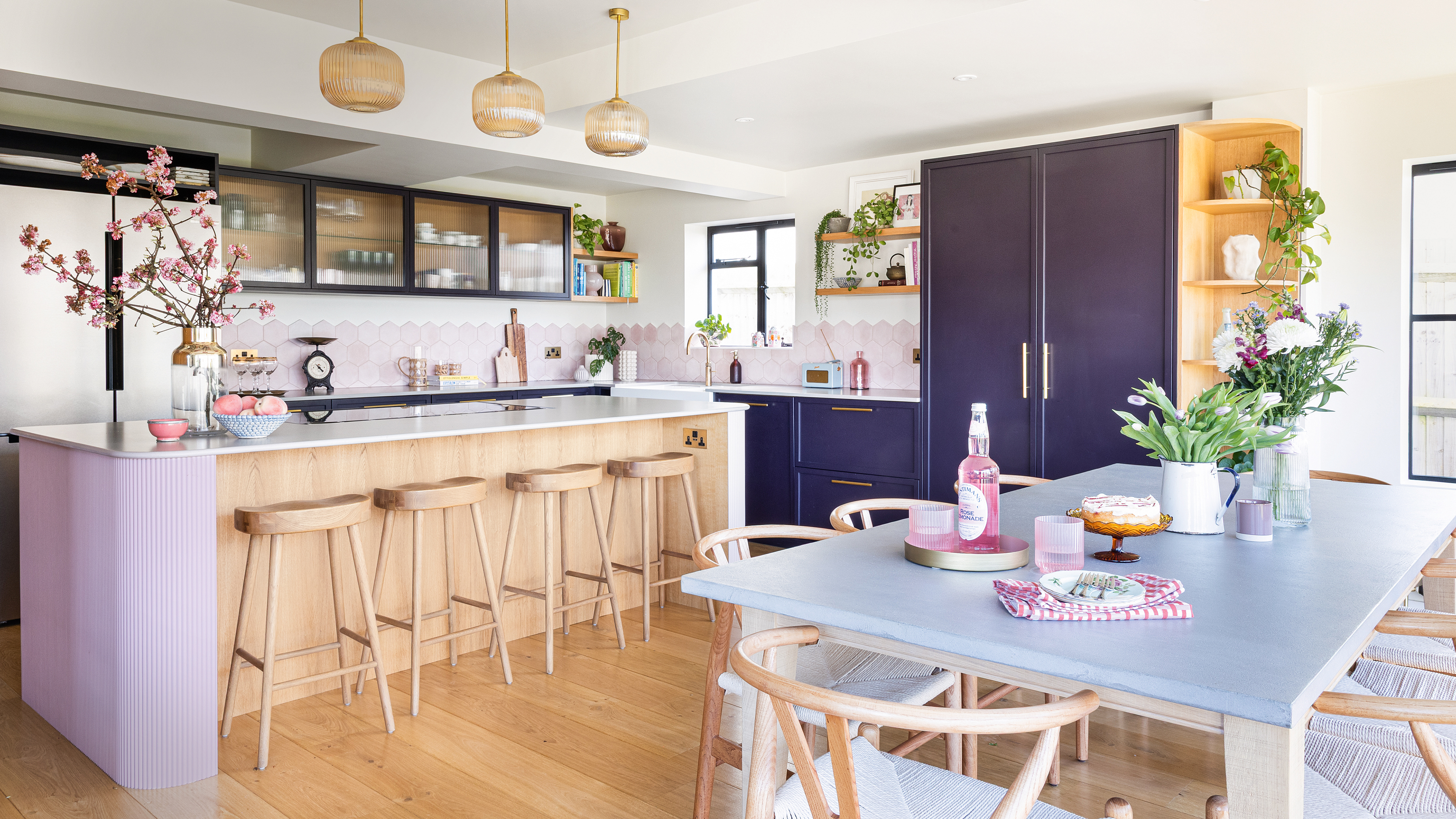 a purple and pink kitchen with purple cabinets and a pale pink hexagonal tiled splashback, pink island and a wooden dining. table and chairs