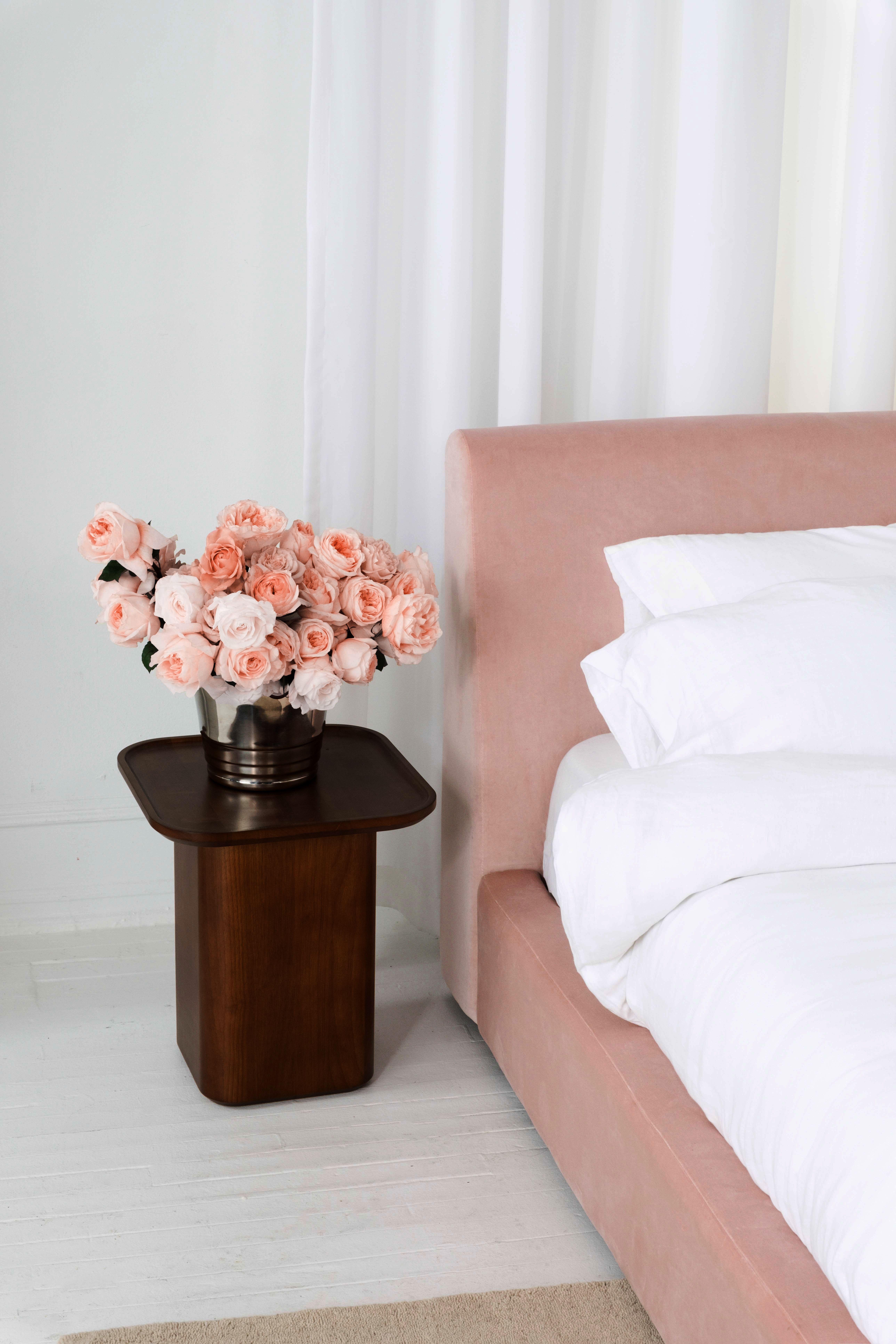 A soft pink bed uphulstored in velvet next to a wooden bedside table with pink roses.