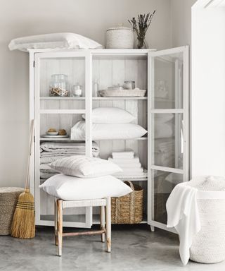 white freestanding linen cupboard with pillows, bedding, baskets, glass containers, stool with pillows, broom