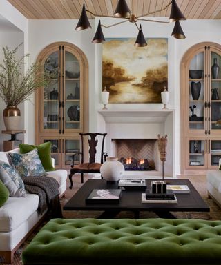 white living room with green footstool, green cushions, white couch, black wood coffee table, alcove shelving, shiplap ceiling, retro metal pendant light, artwork, artisanal pieces,