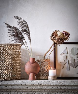 Mantel decorated with lit candle, woven basket, vase of dried flower stems, frame with geometric cut out collage