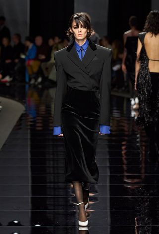 how to wear velvet is shown in a photo from Sportmax's fall 2024 runway show with a model wearing a blue button-down shirt under a black blazer tucked into a black velvet midi skirt with sheer tights and white pumps