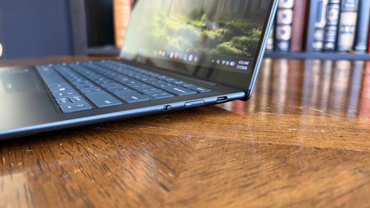 A sleek laptop on a wooden table, showing side view with USB-C ports and keyboard. The screen displays a blurred forest image, creating a modern and serene vibe.