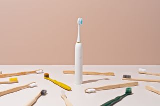 Electric toothbrush standing upright surrounded by regular toothbrushes in front of a beige background