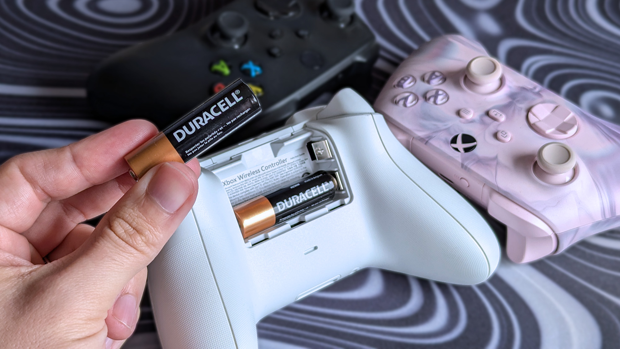 Holding Duracell AA battery with various Xbox Series X|S controllers on a patterned desk