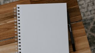 En blank anteckningsbok ligger på ett träfärgat bord bredvid en penna.