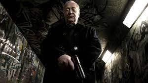 A screenshot of Harry Brown looking down into the camera as he holds a pistol in his self-titled film