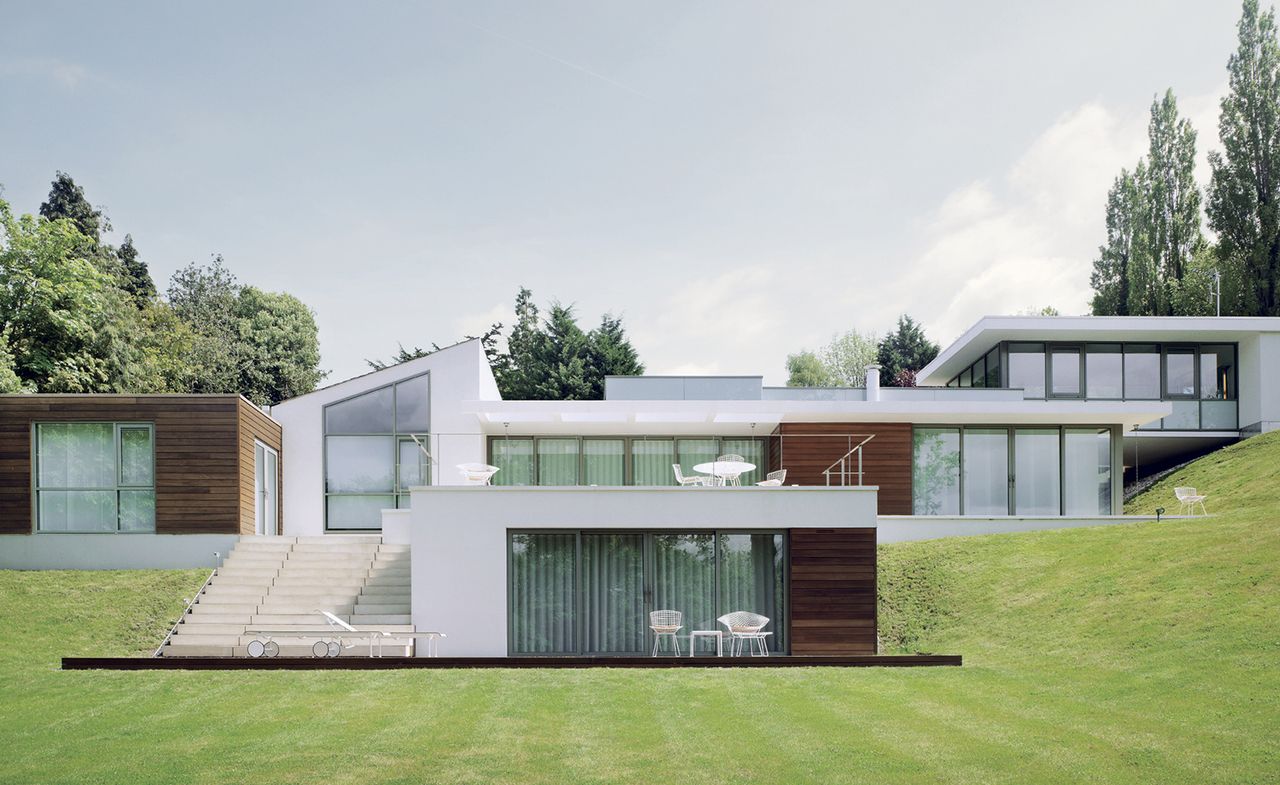 Exterior view of Gerry McGovern&#039;s white and wood panelled Warwickshire multi-level home and surrounding greenery during the day