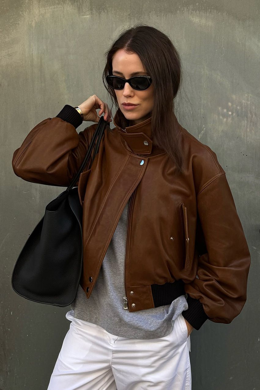 A woman standing outside wearing a brown funnel neck bomber jacket styled with a gray t-shirt, white jeans, a large black tote bag, black sunglasses, and a two-tone watch.