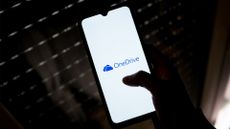 Microsoft OneDrive logo and branding pictured on a smartphone screen held in a human hand while hovering over a laptop keyboard.