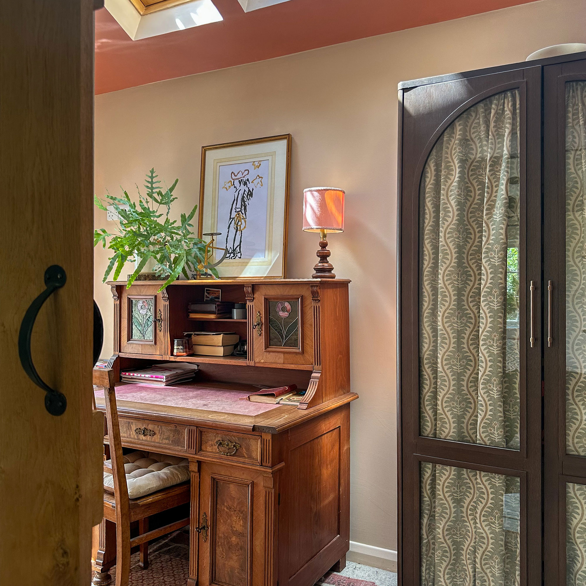 Home office with vintage wooden desk and dark wooden wardrobe with glass doors backed with green patterned fabric