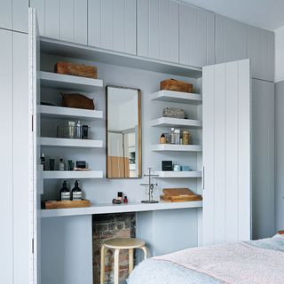 A bedroom with large cupboard doors open to show a dressing table within, woodwork painted pale grey with built in wall of cupboards and storage
