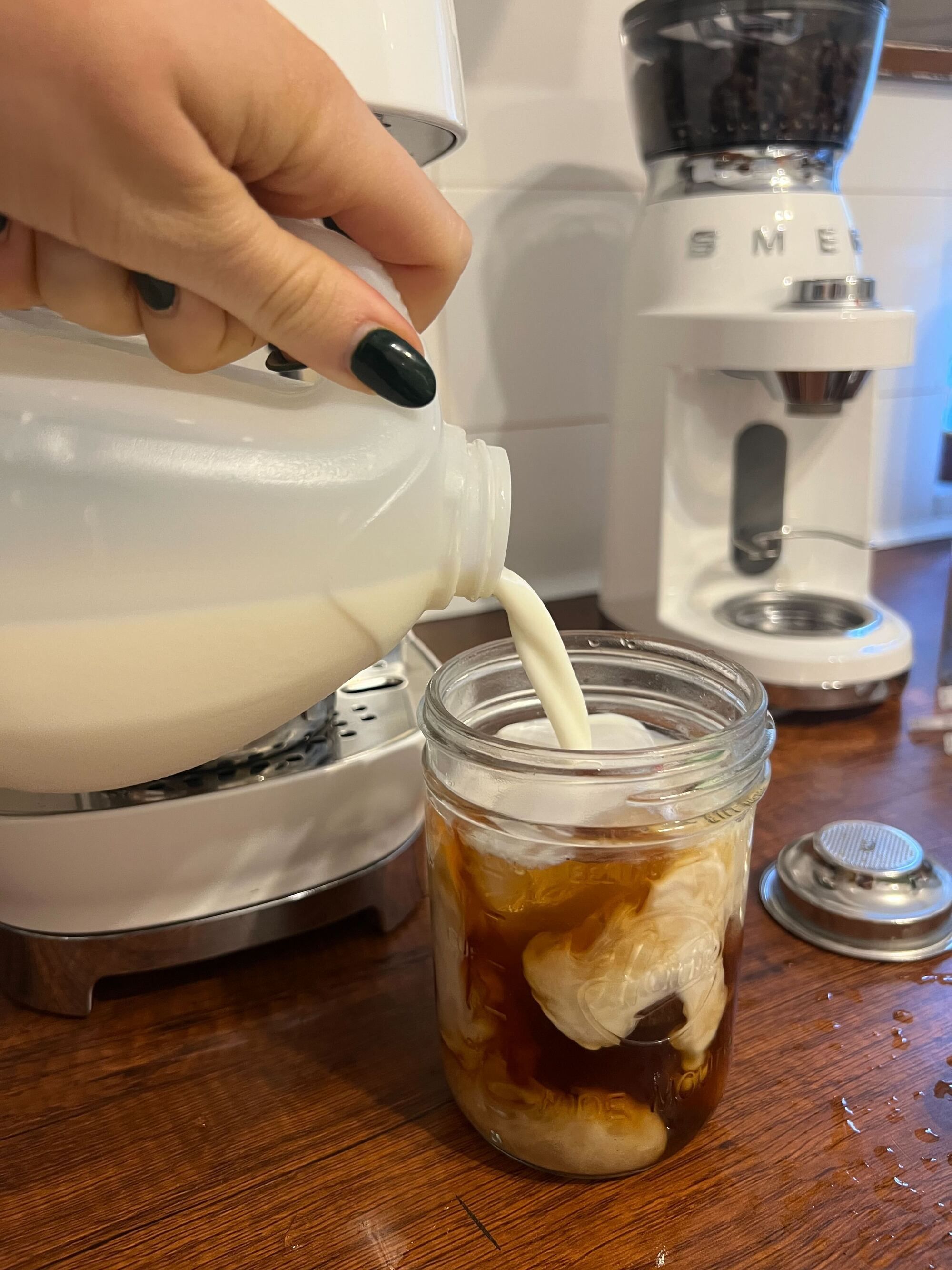 Image of a hand pouring milk from a jug into a mason jar with cold brew on a wooden countertop. There is a white Smeg cold brew machine and coffee grinder in the background.