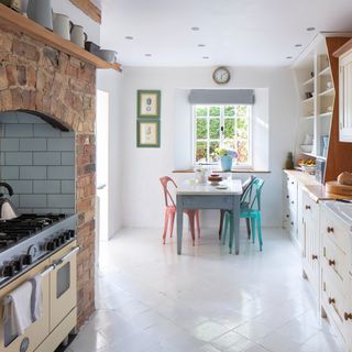 narrow white kitchen with colourful cafe chairs, cream range in brick fireplace and cream units