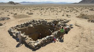 Archaeologists examine a large square-shaped stone burial in the desert.