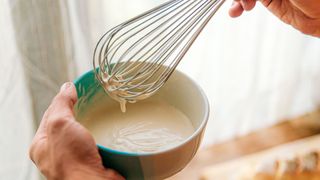 A hand holding a bowl of batter and a handheld whisk, a little batter dropping off it