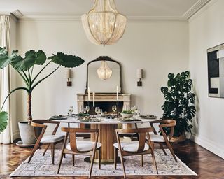 Luxury cream dining space with round dining table, patterned rug, large houseplants, and ornate mirror on mantel.