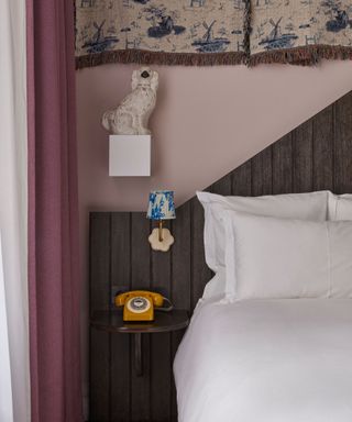 pink purple hotel bedroom with a panelled headboard, small wall sconce, a plinth with a vintage beswick dog and wall tapestry hangings above