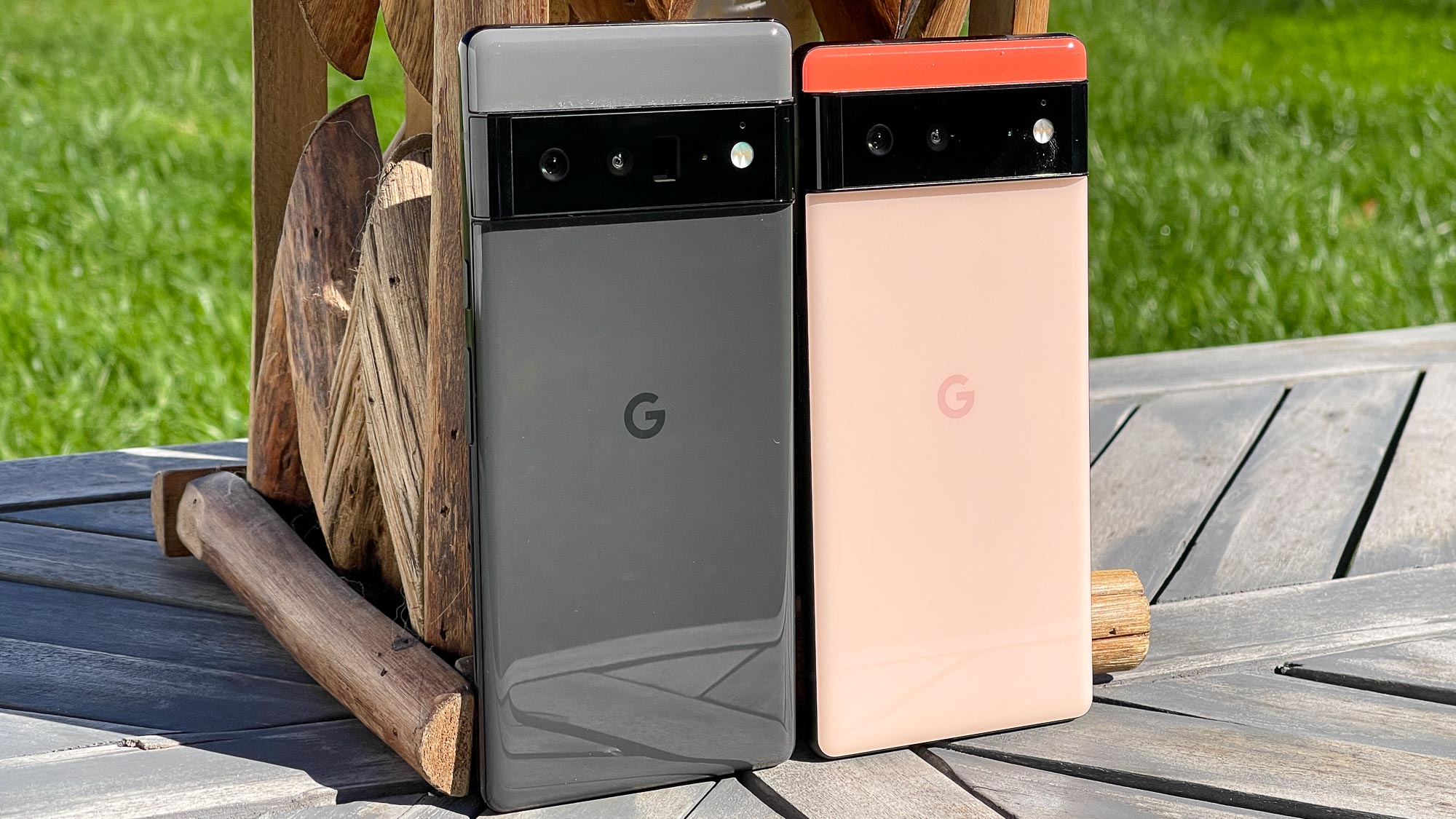 The Google 6 Pro (left, in black) and Google Pixel 6 (right, in coral), leaning together against a tree on wooden decking