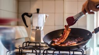 Cropped Hand Cooking Bacon On Stove In Kitchen
