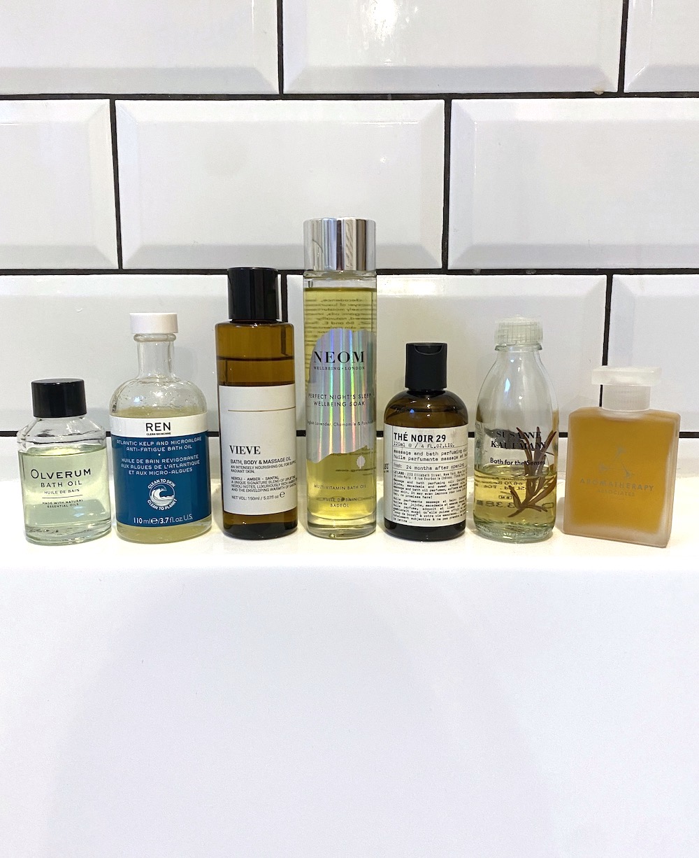 A group shot of some of the best bath oils featured in this guide from (left-right) Olverum, REN, VIEVE, NEOM, Le Labo, Susanne Kauffmann and Aromatherapy Associates, stood on the edge of a bath in front of a white tiled background