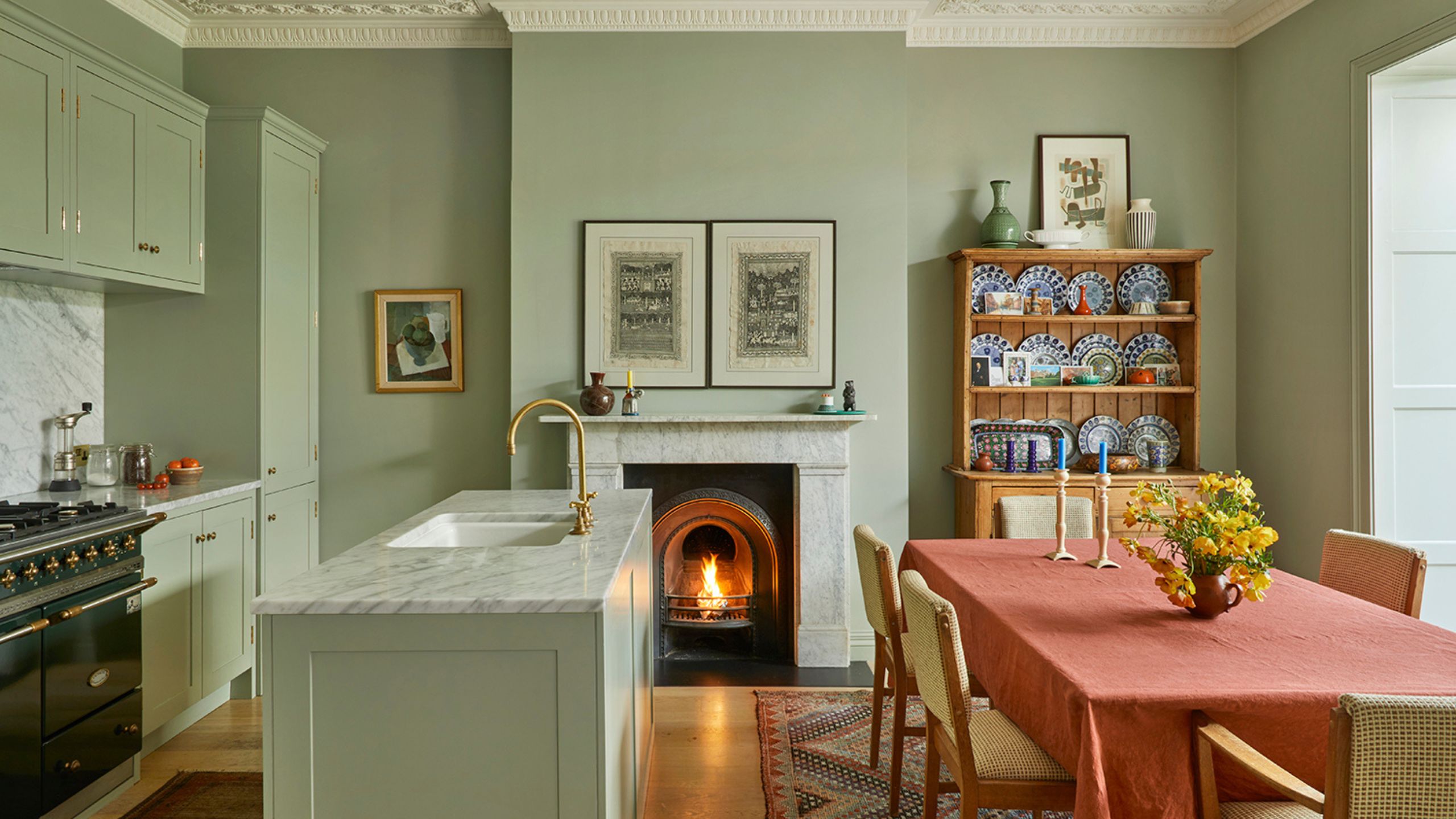 Kitchen painted mint green, a dining table with an orange tablecloth, a marble island, pale green cabinets, and a large fireplace at the centre.