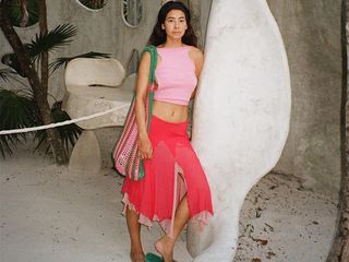 Michelle Li leans on building pillar while wearing a pink tank top, a red midi skirt, and green flip-flops. She is carrying a large crochet shoulder bag.