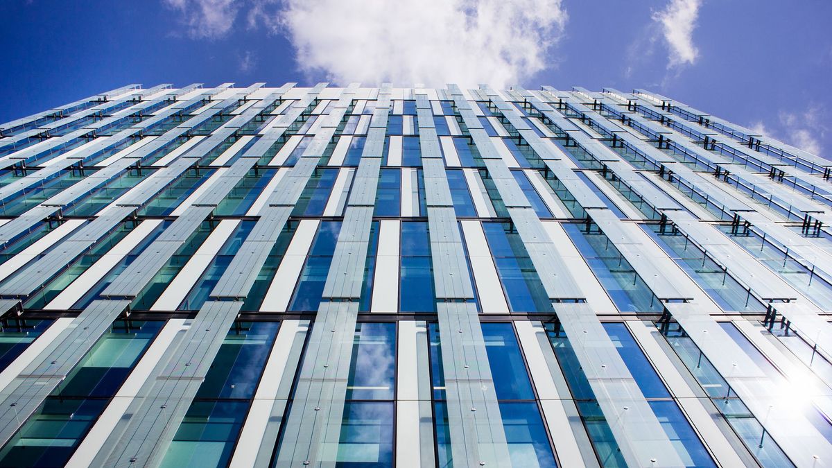 A close up photo of a building angled towards a cloudy blue sky, showing white and grey panels next to blue windows