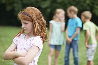 A little girl stands looking sad while other children gossip about her.