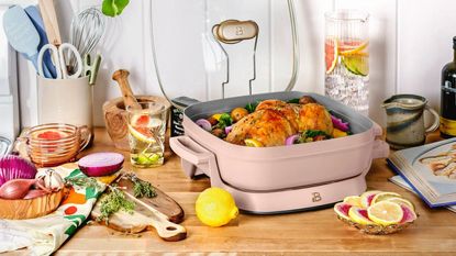 Pale pink electric skillet on wooden countertop next to chopping boards and vegetables