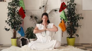 Guru Jagat, sitting cross-legged with her hands pressed together as she sits in front of plants and colored flags, in 'Breath of Fire.'