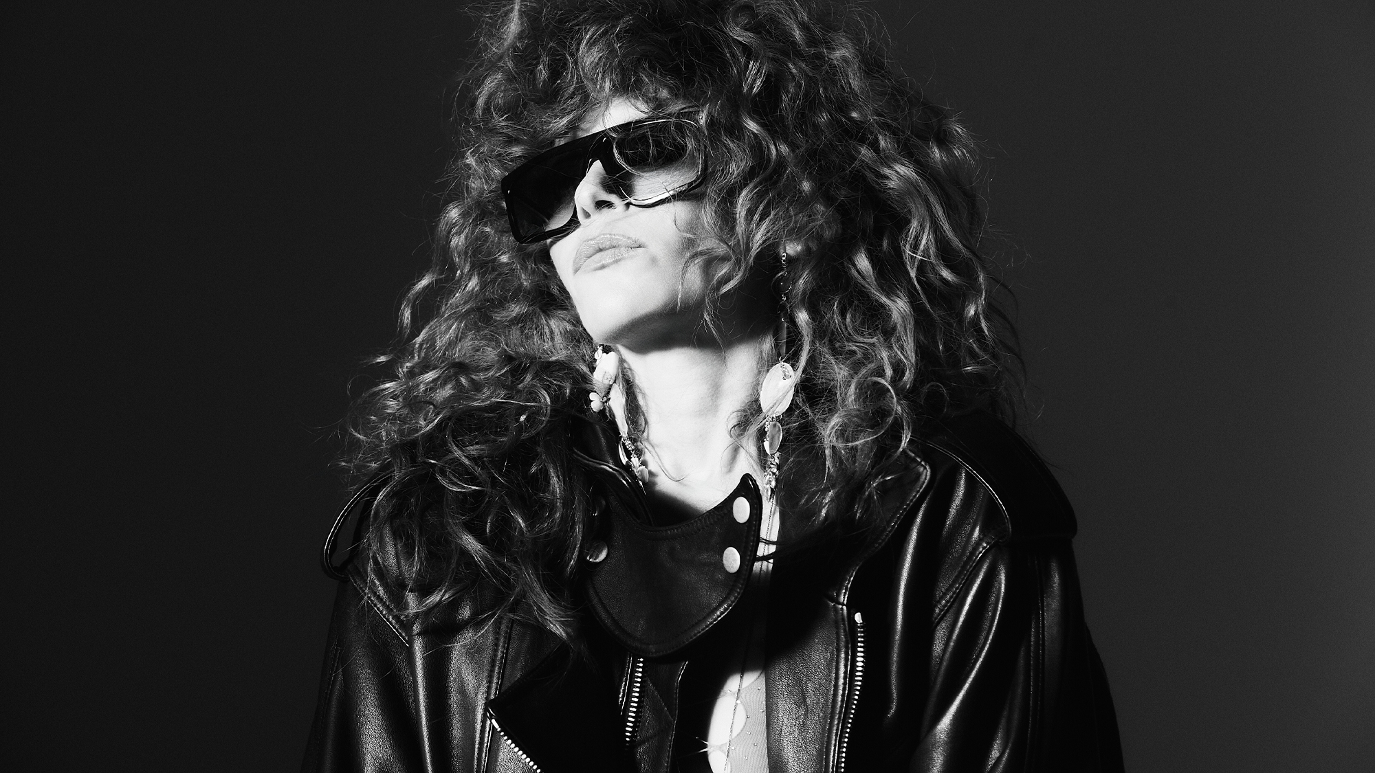 Natasha Lyonne shot in black and white by Andrew Arthur. She is wearing oversize sunglasses, drop earrings, and a black leather jacket. 