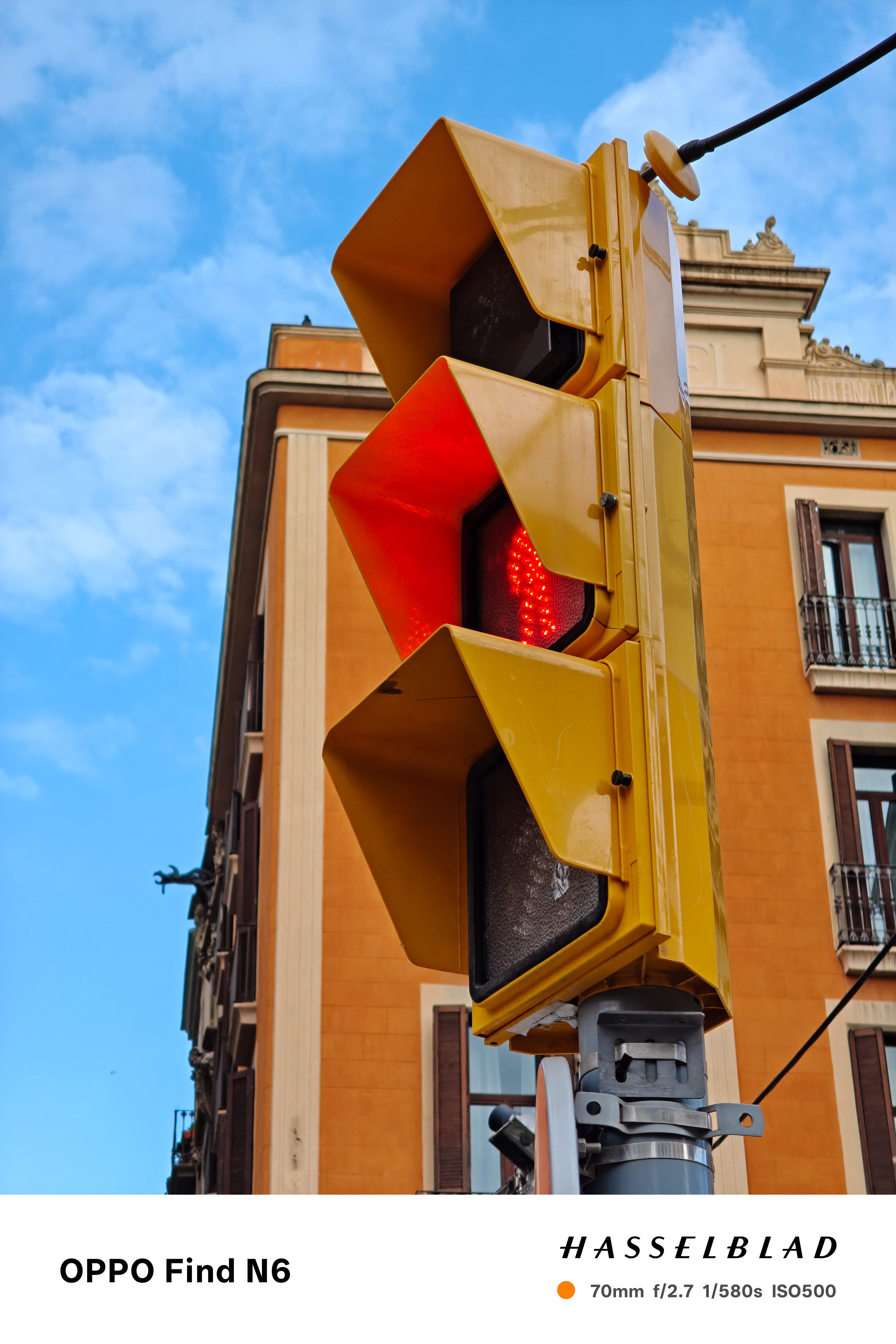 A low-angle, close-up shot of a bright yellow traffic light housing. The middle light is illuminated with a red "don't walk" hand symbol. In the background, an orange Mediterranean-style building with dark shutters is visible under a slightly cloudy blue sky.