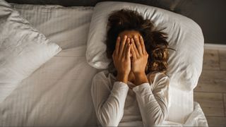 Woman wearing white pyjamas holding her hands over face as she lays on her back in bed.