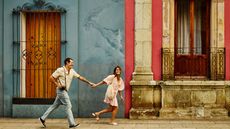 A couple in their 40s runs playfully down a street in Oaxaca, Mexico. He carries a camera.