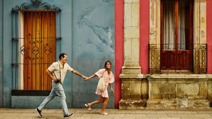 A couple in their 40s runs playfully down a street in Oaxaca, Mexico. He carries a camera.