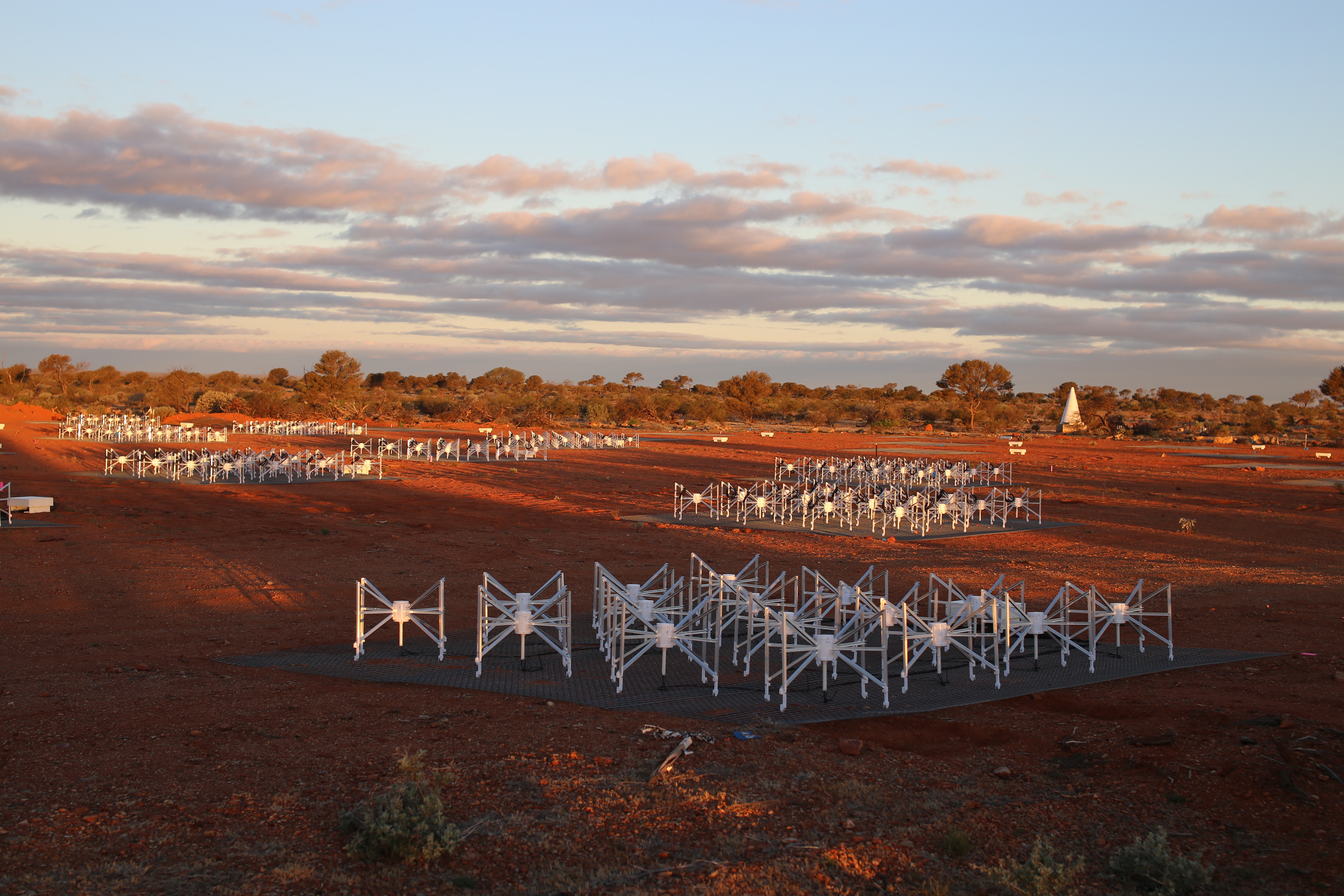 A red-brown field stretches to a horizon of low trees beneath a blue cloudy sky. White, spider like contraptions stand in groups on grey mats around the field.