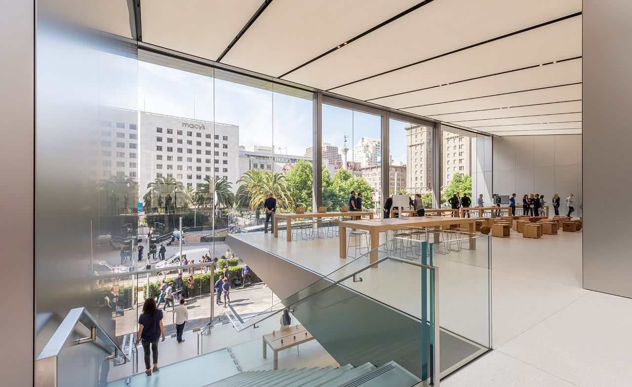 Interior view of the upper level at Apple&#039;s San Francisco store featuring light coloured flooring, long wooden tables with products on top, stools, large windows and a staircase with a metal and glass banister. There are multiple people in the store