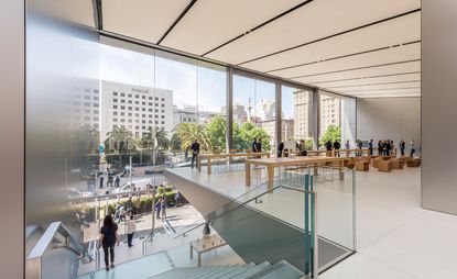 Interior view of the upper level at Apple's San Francisco store featuring light coloured flooring, long wooden tables with products on top, stools, large windows and a staircase with a metal and glass banister. There are multiple people in the store