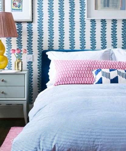 pink and blue bedroom with striped wallpaper and bedding and large yellow bedside lamp with a gold vase and pink peonies on a sage green bedside table