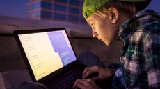 Youth hacker concept image showing teenage boy coding on a laptop in a dimly-lit room.
