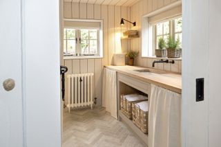 Narrow utility room ideas showing herringbone flooring, wooden worktops and fabric drapes to hide items stored under the sink