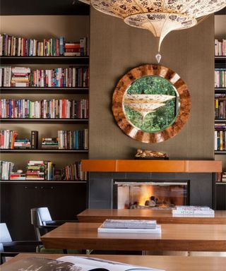 A living room with black bookshelves with colorful books, a domed chandelier, a black fireplace, and two wooden desks with black chairs in front and white books on top