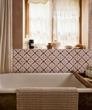 A bathroom with brown washed walls, a window with white curtains, white and dark brown patterned tiles, and a bath with a wooden bath tray and a beige towel hanging from the edge of it