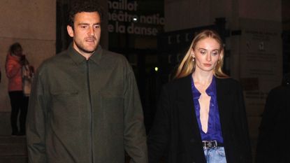Sophie Turner wearing an open blue button down with a black wool coat and light wash jeans 